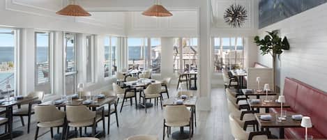 A spacious dining area with multiple tables and chairs, large windows offering a view of the sea, and hanging pendant lights.