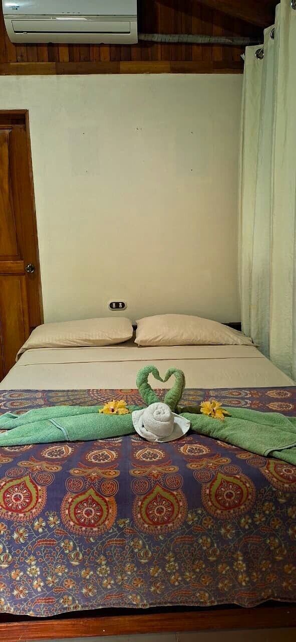 Een bed met een bedspreider met een patroon, een hartvormige decoratie op het bed, en een luchtverkoeler aan de muur.