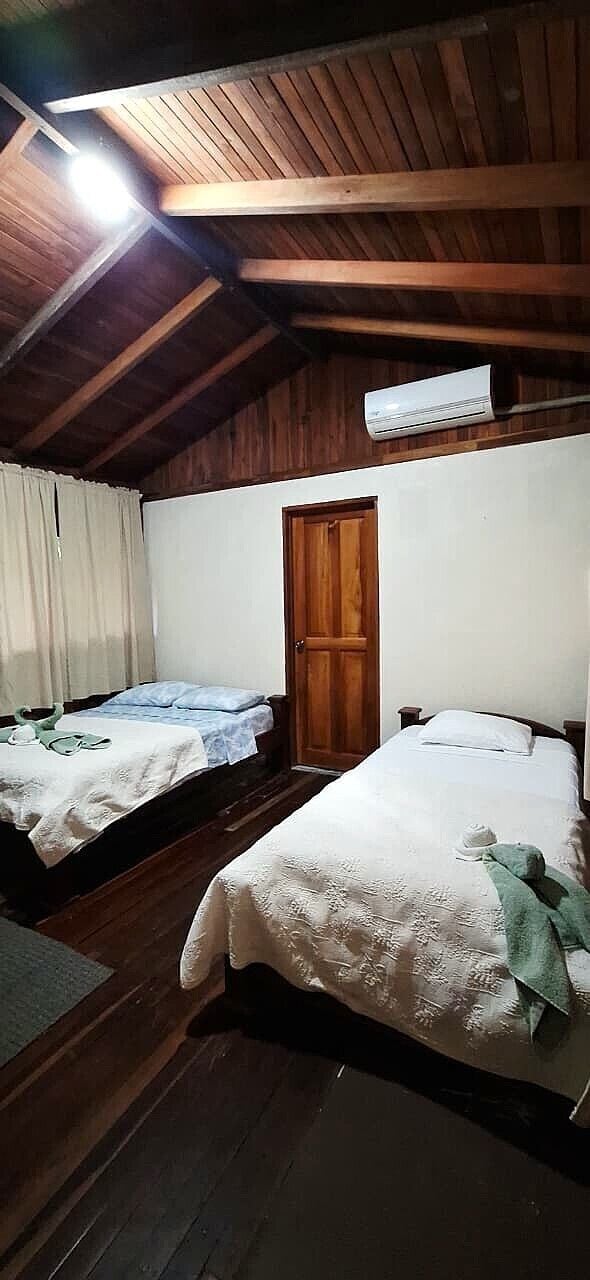 Quarto com duas camas, piso de madeira, teto de madeira, uma porta e uma luminária no teto.