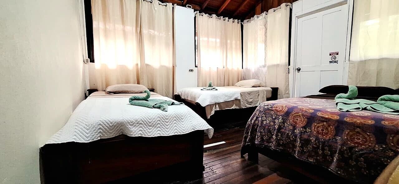 A room with three single beds, each with a different patterned bedspread. There are curtains on the windows, and a white door with a sign on it.