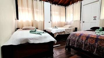 A room with three single beds, each with a different patterned bedspread. There are curtains on the windows, and a white door with a sign on it.