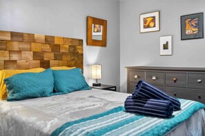Een hotelkamer met een houten hoofdbord, een bed met blauwe en gele beddengoed, een nachtkastje met een lamp en een kledingkast met lades.