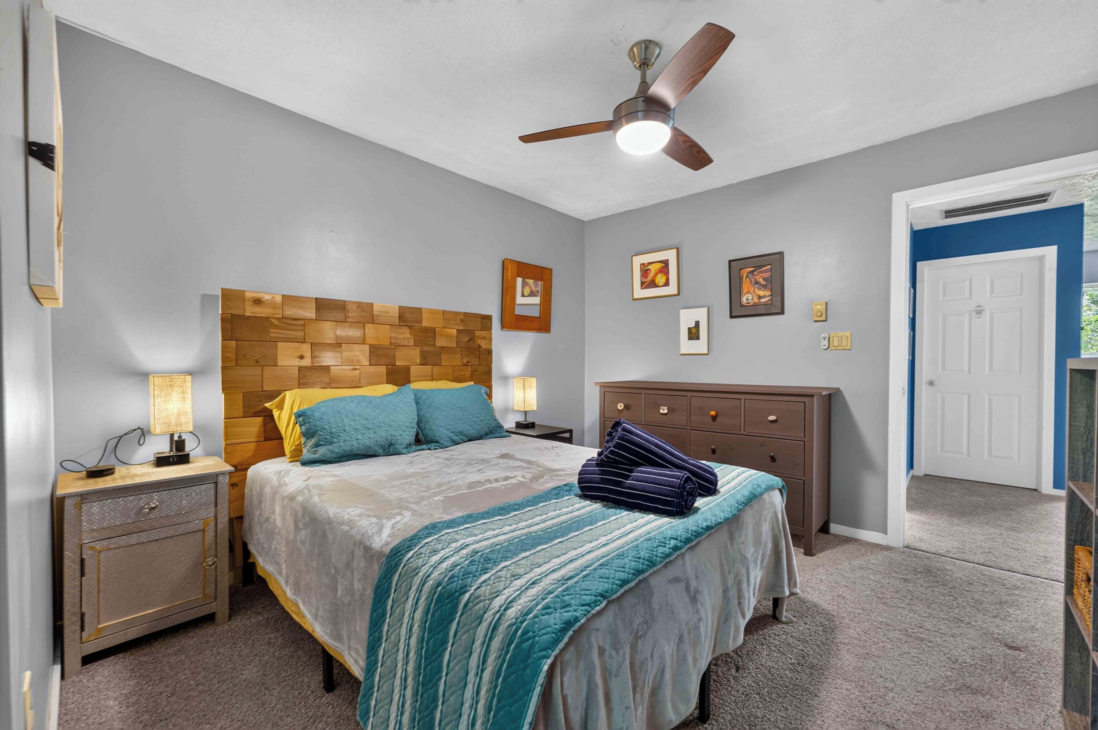 A bedroom with a wooden headboard, a bed with a striped blanket, a nightstand with a lamp, a dresser, and a ceiling fan.