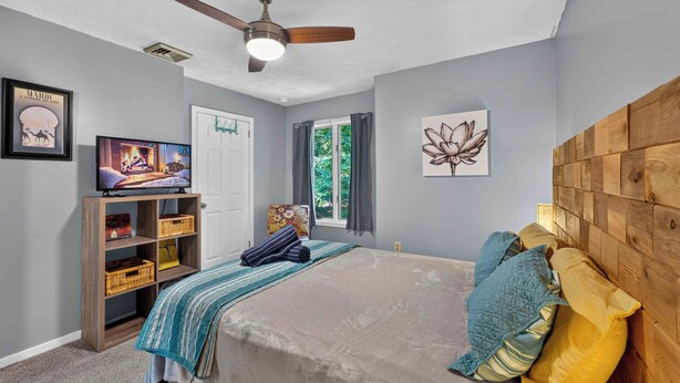 Une chambre à coucher avec un lit, un ventilateur de plafond, un téléviseur posé sur une étagère en bois et des œuvres d’art accrochées aux murs.