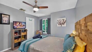 A bedroom with a bed, a ceiling fan, a TV on a wooden shelf, and artwork on the walls.