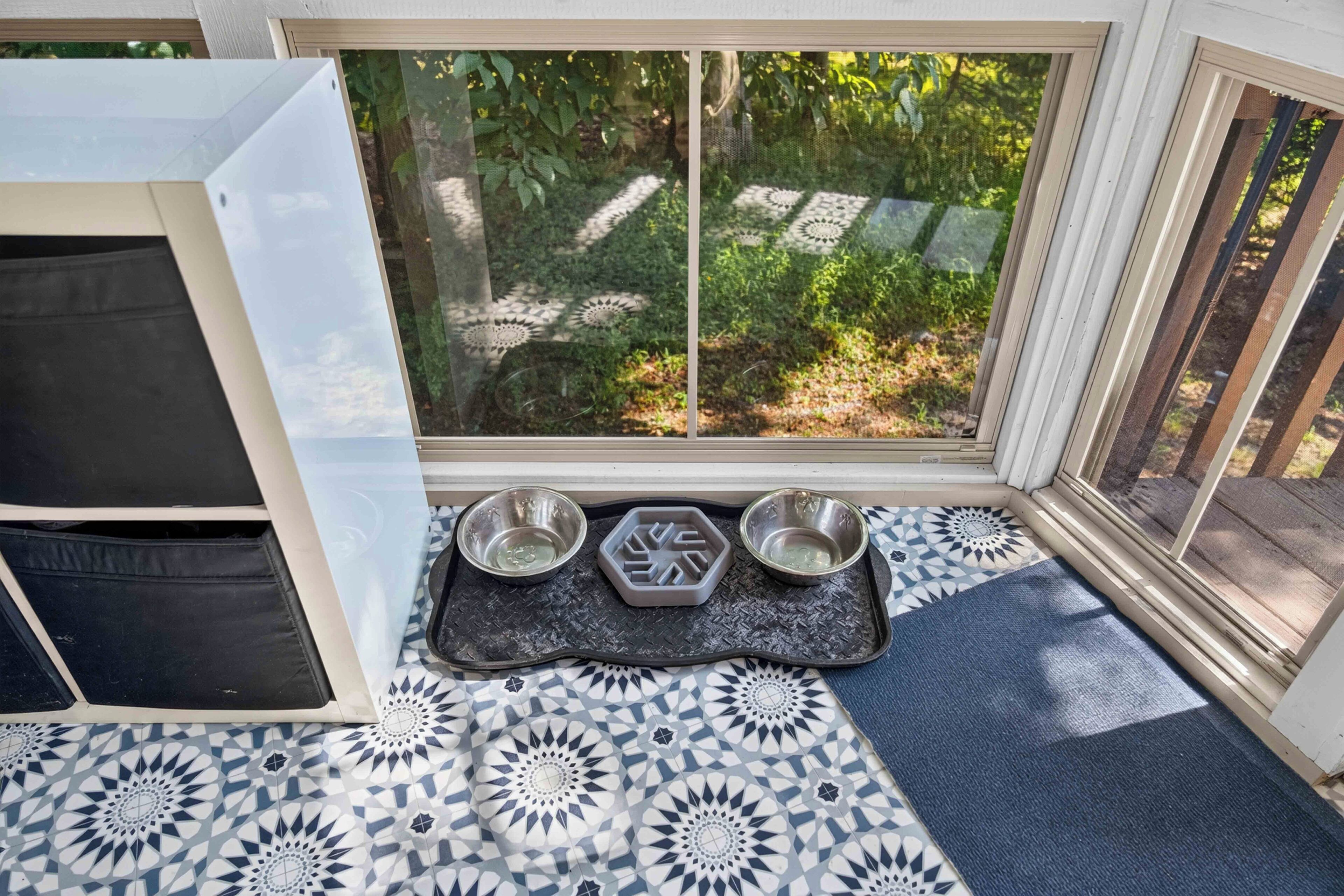 Un espace dédié à l’alimentation des animaux de compagnie, avec deux gamelles et un distributeur d’eau posés sur un tapis, un sol à motifs et une fenêtre donnant sur un paysage verdoyant.