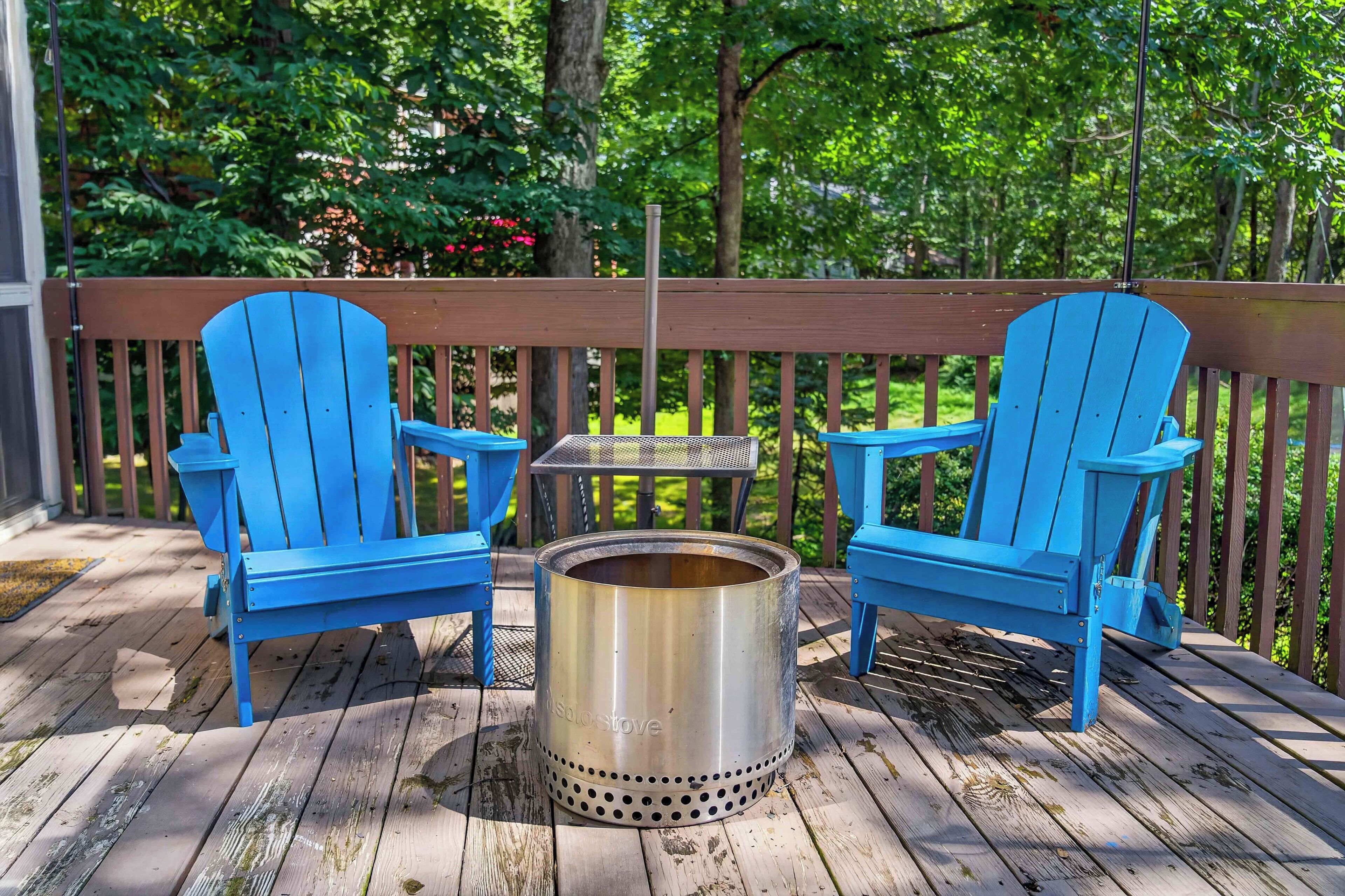 A wooden deck with two blue Adirondack chairs and a metal stove.