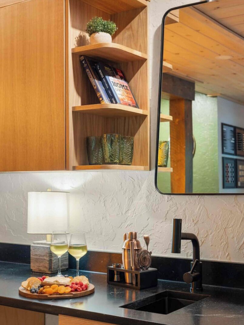A kitchen with a wooden shelf, a mirror, a countertop with a glass of wine, and a fruit platter.