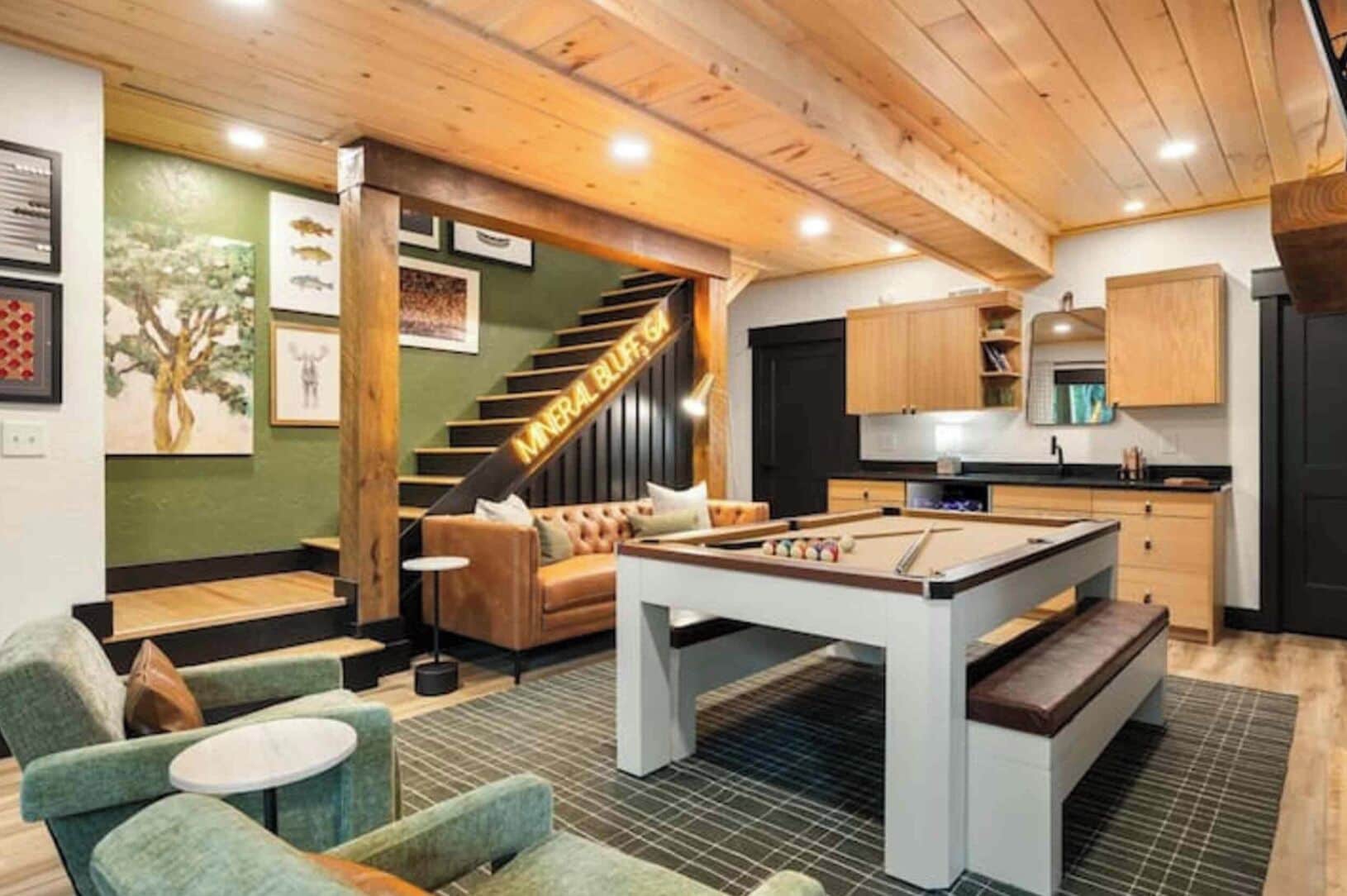 A cozy room with a pool table, green upholstered chairs, a staircase, and a kitchen area.