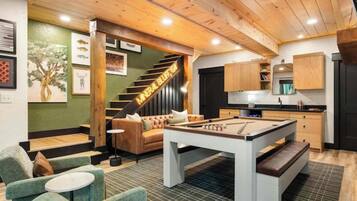 A cozy room with a pool table, green upholstered chairs, a staircase, and a kitchen area.