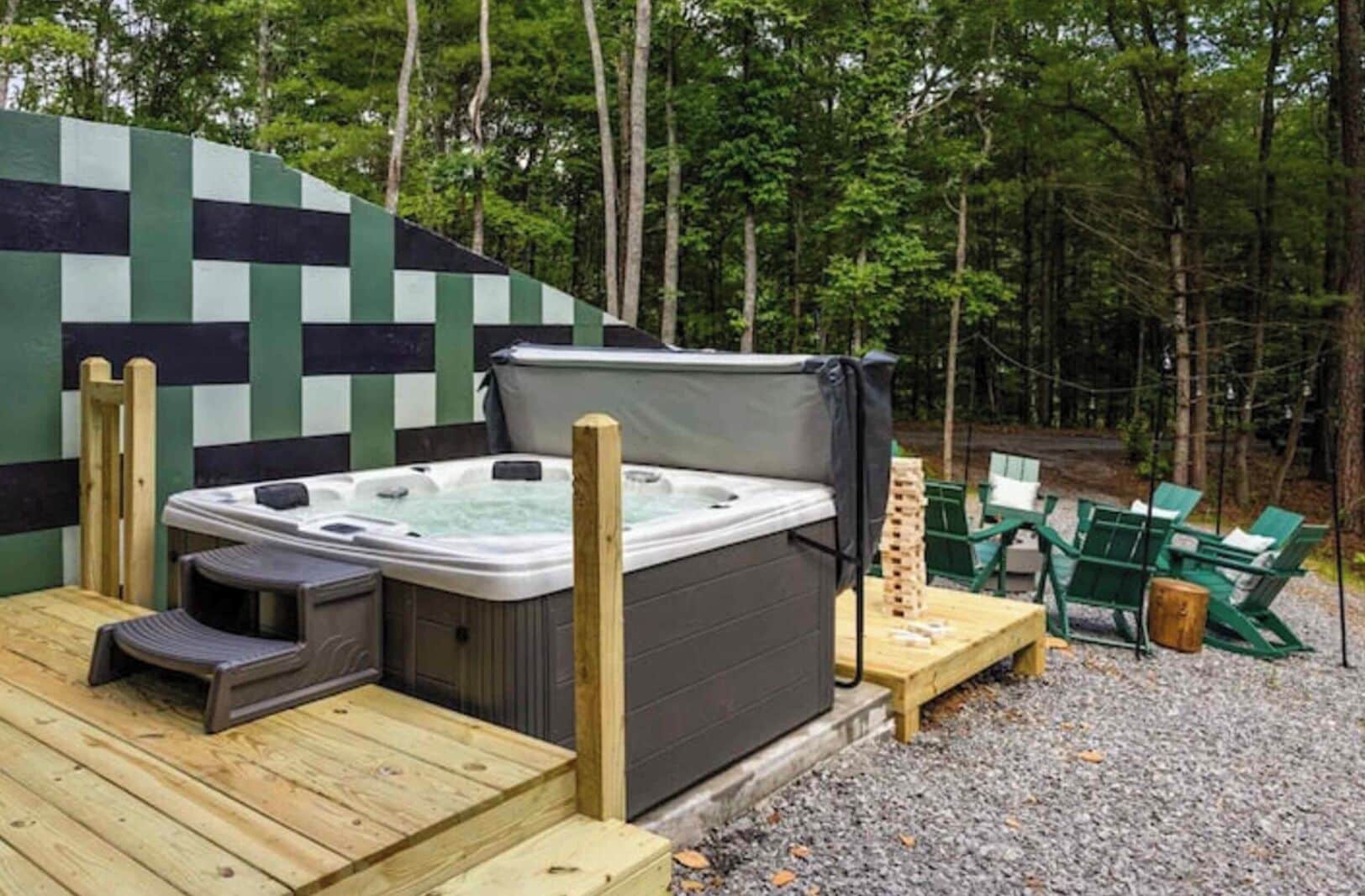 Jacuzzi en terraza de madera con fondo de pared a cuadros, rodeado de mobiliario exterior y zona boscosa.