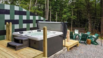Jacuzzi en terraza de madera con fondo de pared a cuadros, rodeado de mobiliario exterior y zona boscosa.