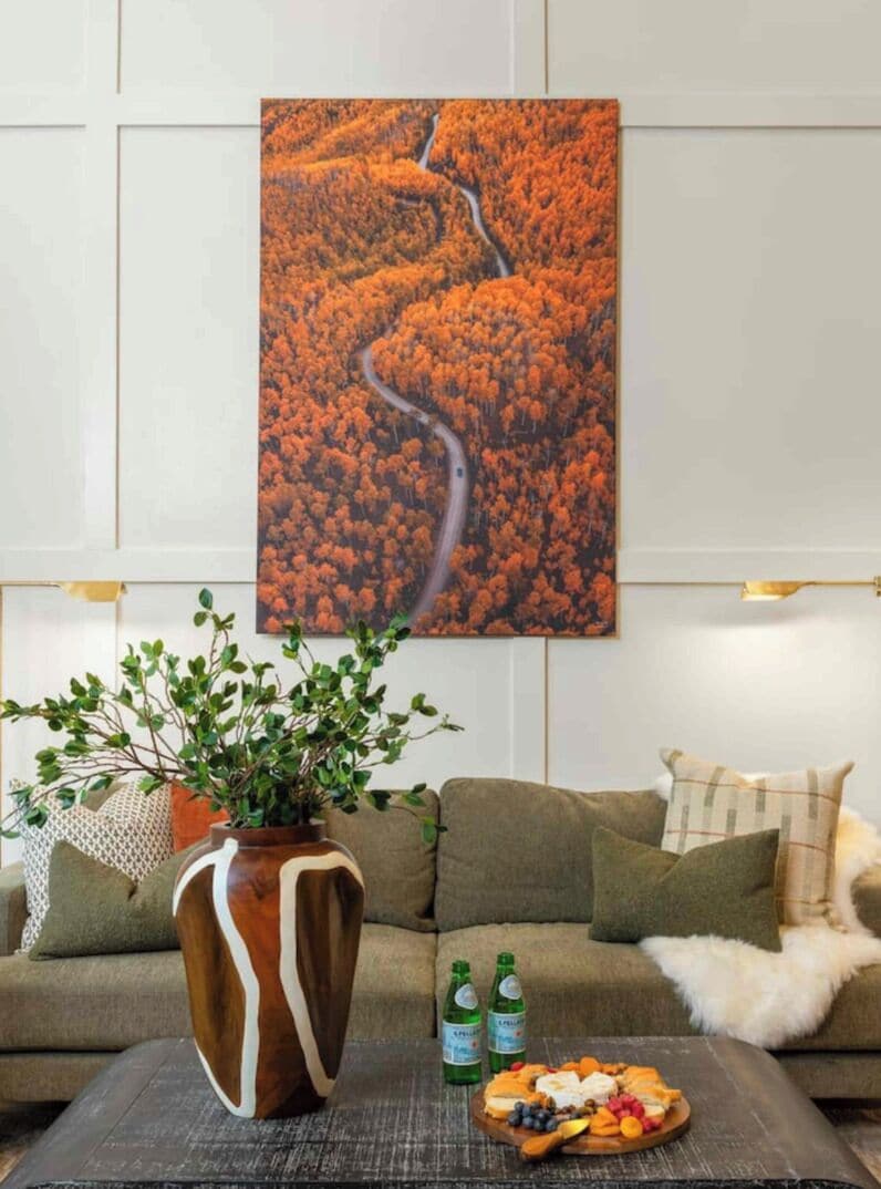 A living room with a sofa, a coffee table with a fruit platter, a vase with green foliage, and a painting of a winding road through autumn foliage.