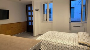 A bedroom with a bed, a wooden headboard, a TV mounted on the wall, and a window with a view of the outside. - Apartment in Downtown Chalamont (Chalamont)