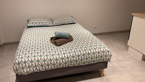 A single bed with a patterned bedspread and pillows, a rolled-up towel, and a wooden nightstand to the right. - Apartment in Downtown Chalamont (Chalamont)