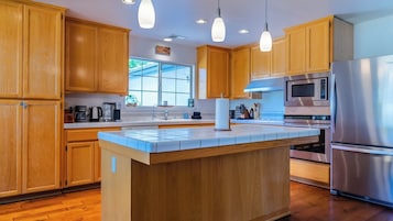 Cocina moderna con gabinetes de madera, una isla central, electrodomésticos de acero inoxidable, iluminación colgante y un ventanal que proporciona luz natural.
