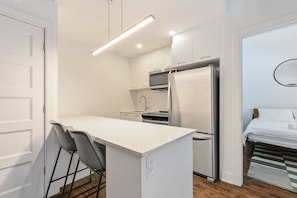 A compact kitchen with a white countertop, a refrigerator, and a stove. There is a small seating area with a chair and a table. - Modern 1BR • Walk to Old Port (Montreal)