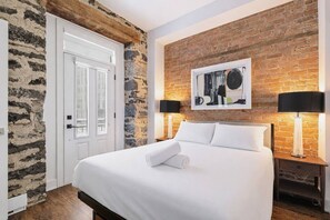 A hotel room with a bed, a stone wall, a brick wall, a door, and two bedside lamps. - Modern Downtown Montreal 2-Bedroom • Steps to Old Port (Montreal)