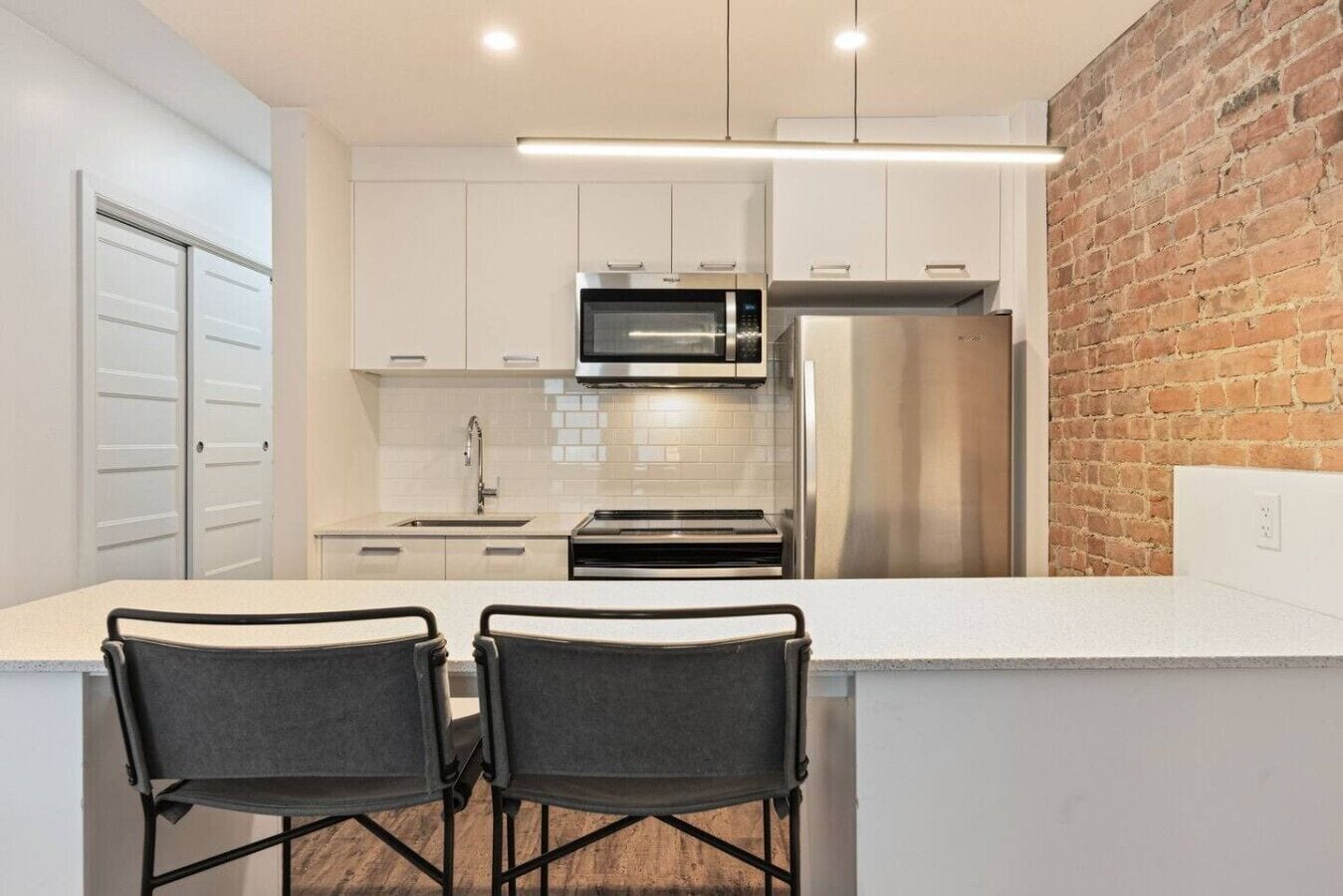 A modern kitchen with white cabinets, a brick wall, stainless steel appliances, and a bar area with two chairs.