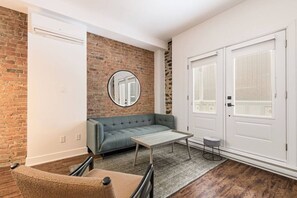 A room with a brick wall, a grey sofa, a round mirror, a white door, and a small stool. - Modern Downtown Montreal 2-Bedroom • Steps to Old Port (Montreal)