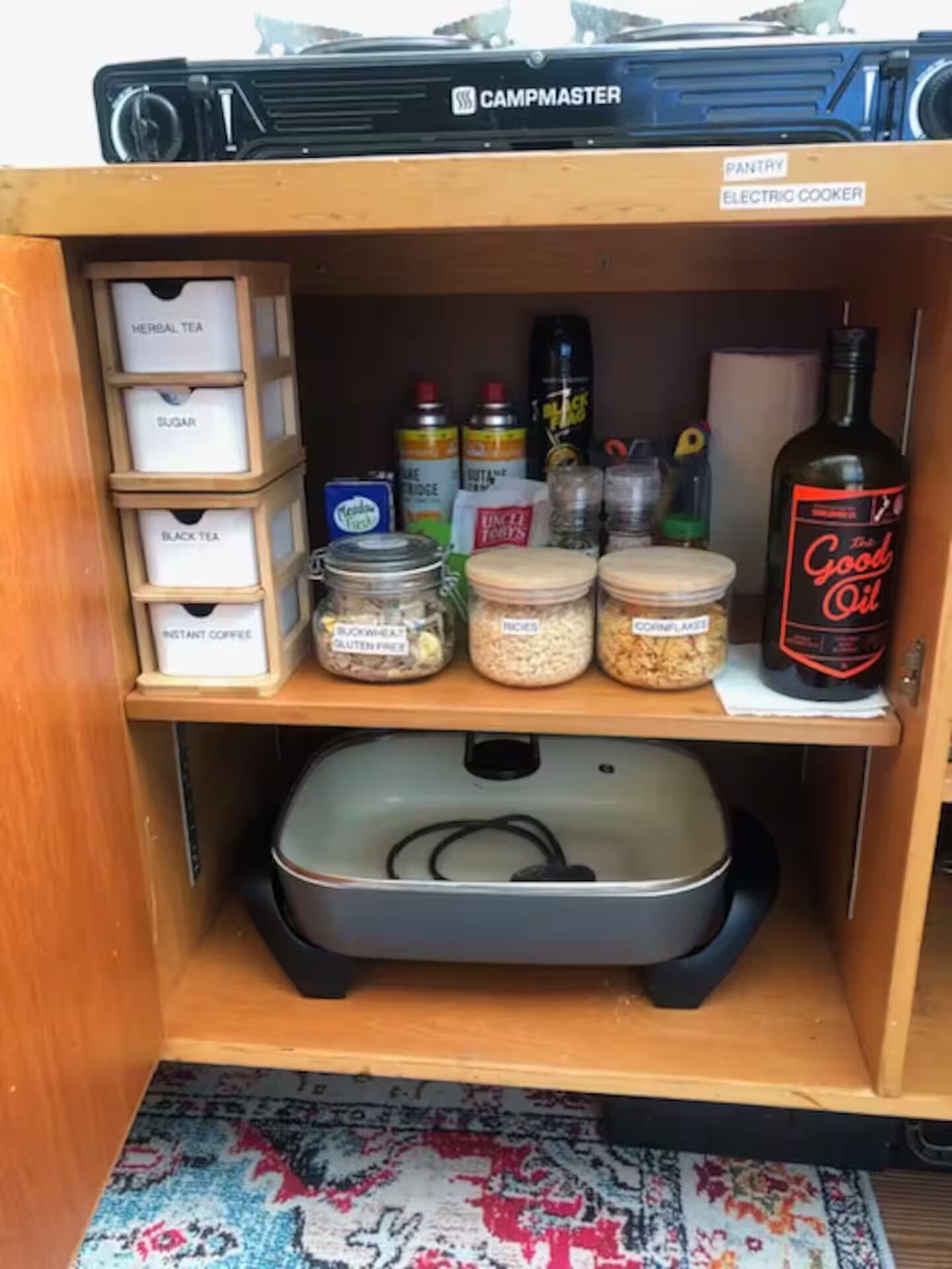 Ein Vorratsschrank mit verschiedenen Lebensmitteln, darunter Zuckergläser, Tee und Instantkaffee, eine Flasche Kochöl und ein CampMaster Elektrokocher.