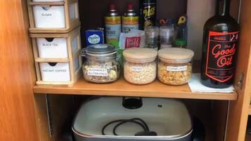 Ein Vorratsschrank mit verschiedenen Lebensmitteln, darunter Zuckergläser, Tee und Instantkaffee, eine Flasche Kochöl und ein CampMaster Elektrokocher.