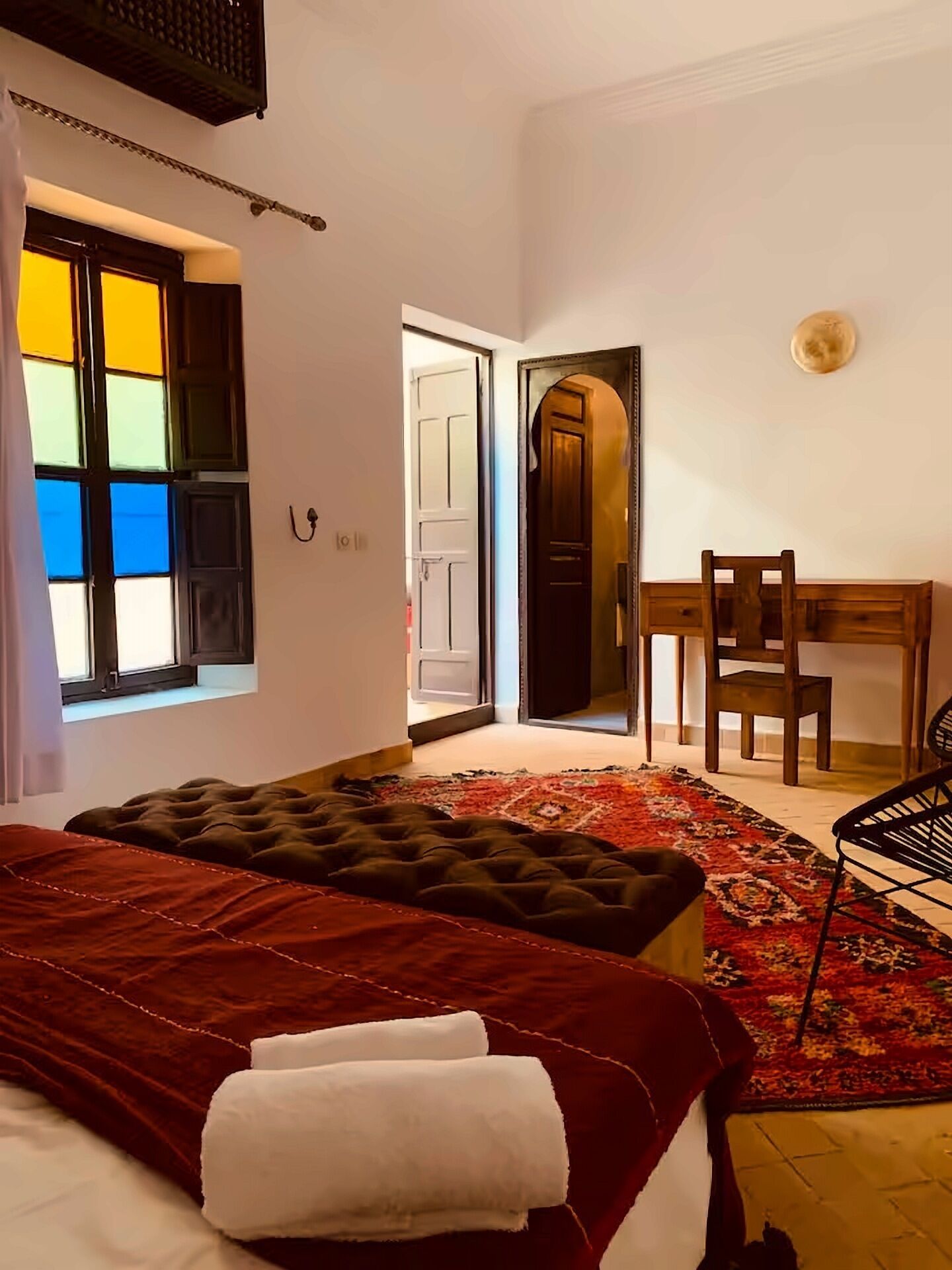 Una camera da letto con un letto, una scrivania e una sedia. Si vede una finestra con persiane colorate e una porta che conduce in un'altra stanza.