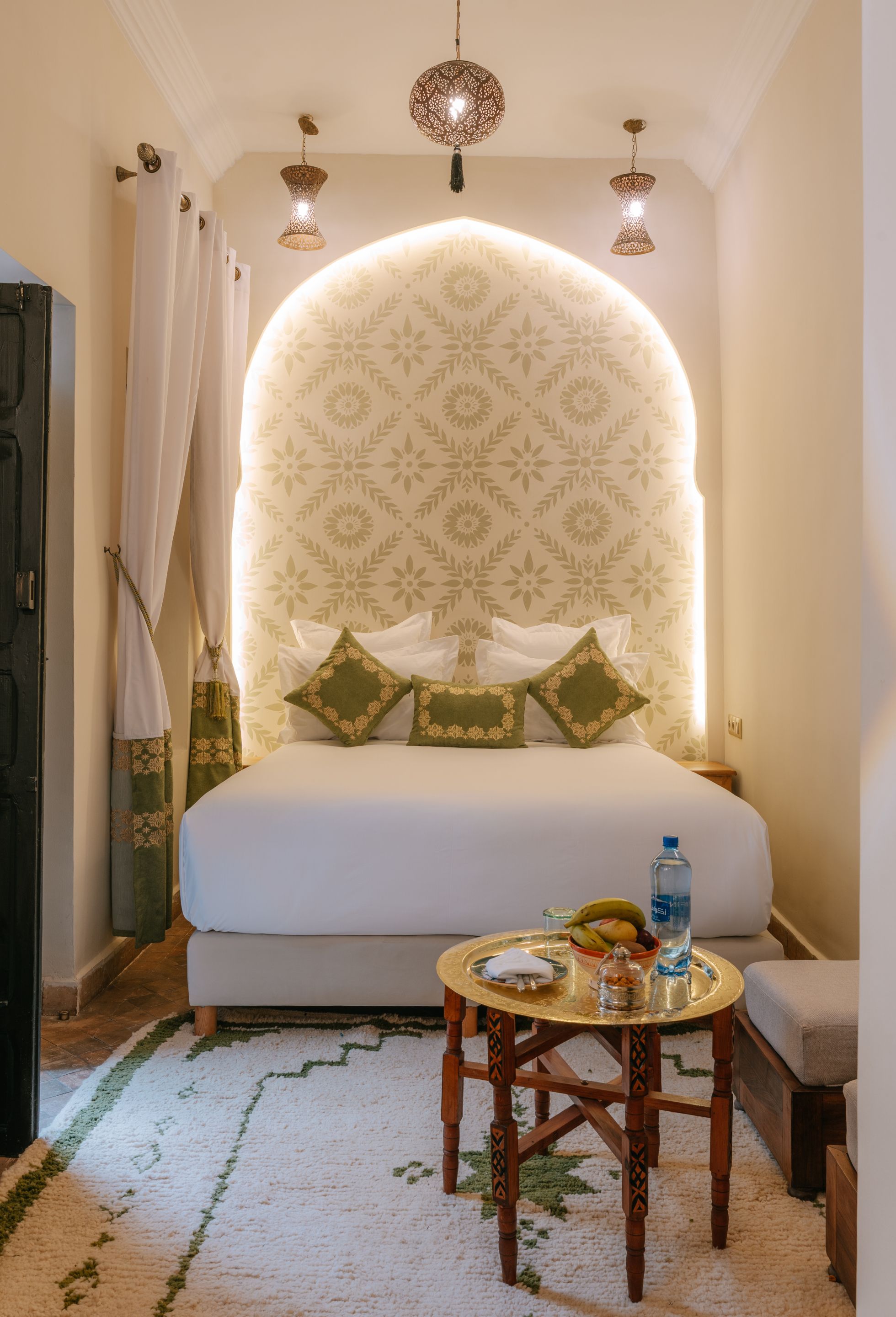Une chambre avec un grand lit, une table de chevet avec une bouteille d’eau et un panier de fruits.