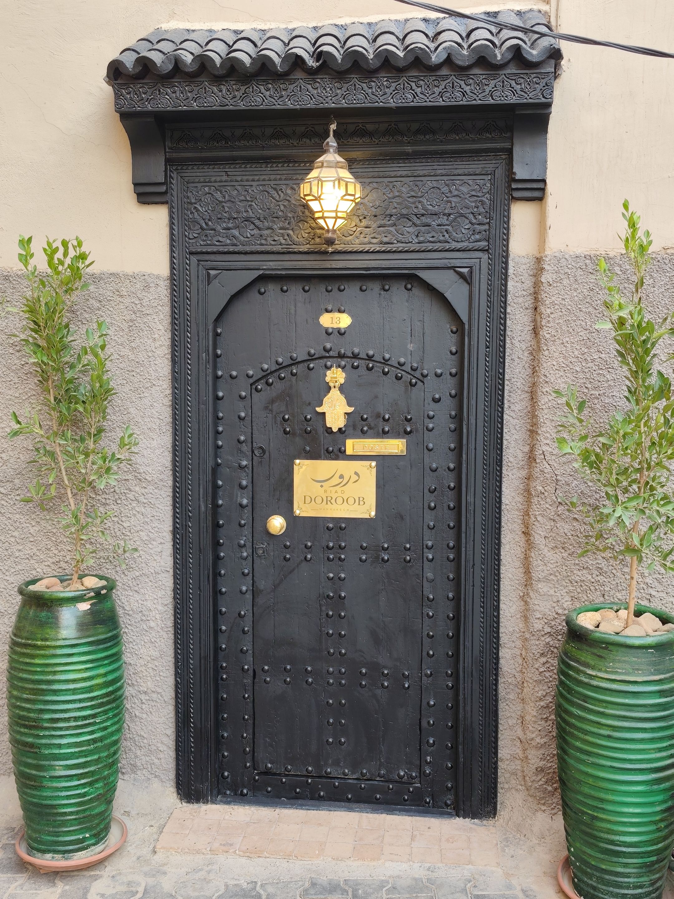 Une porte noire richement décorée, ornée d’une plaque portant un numéro, une lanterne traditionnelle au-dessus, et deux plantes en pot de chaque côté de la porte.