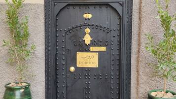 Une porte noire richement décorée, ornée d’une plaque portant un numéro, une lanterne traditionnelle au-dessus, et deux plantes en pot de chaque côté de la porte.