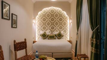 Une chambre d’hôtel avec un lit, une petite table sur laquelle se trouvent une bouteille et des fruits, une chaise et une tête de lit décorative.