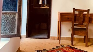 Quarto com escrivaninha de madeira, uma cadeira, tapete estampado e porta que dá para o interior escuro.
