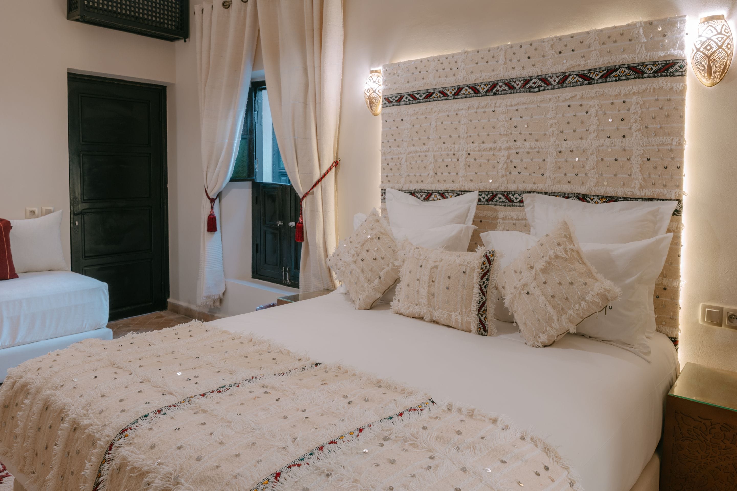 Ein Schlafzimmer mit Bett, Kissen, einem Kopfstück mit Zierleisten, einem Fenster mit Vorhängen und einer Tür.