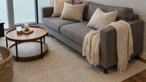 Un salon moderne avec un canapé gris, deux petites tables rondes, une grande fenêtre avec des rideaux, un lampadaire et une œuvre d’art minimaliste accrochée au mur.