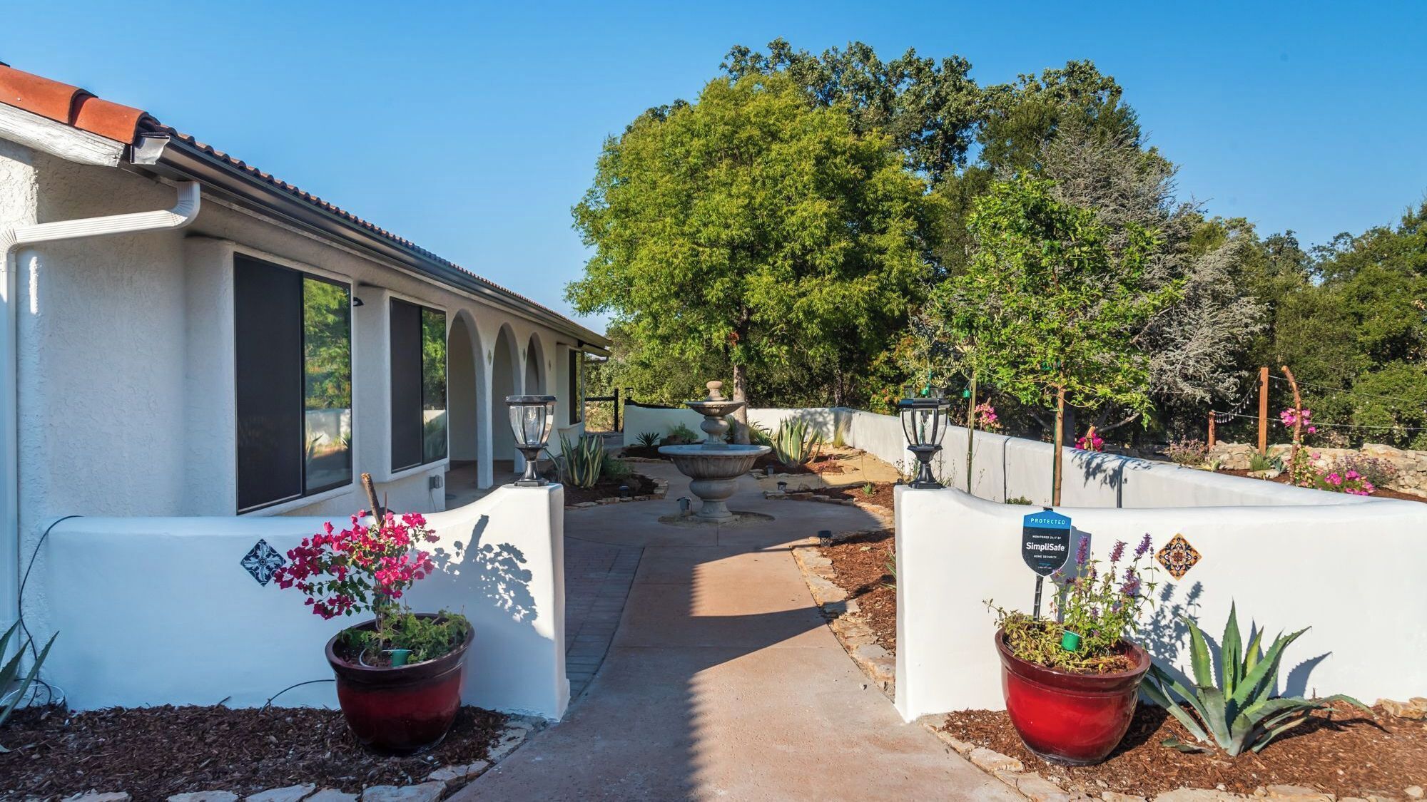 A pathway leading to a building with a tiled roof, surrounded by potted plants and a large tree.