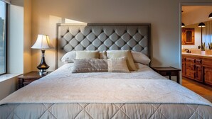 A neatly made bed with a tufted headboard, a bedside lamp, and a view of a wooden kitchen area. - Na Hoku Ranch by Avantstay Spanish Hacienda + Spa + Court 5.5 Acres Near Paso (Atascadero)