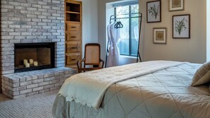 Une chambre avec une cheminée en briques, un lit recouvert d’une couette matelassée, une chaise en bois et, au mur, des reproductions encadrées de gravures botaniques.