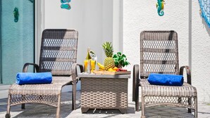 Duas cadeiras de vime com almofadas azuis e uma pequena mesa com frutas e bebidas no centro.