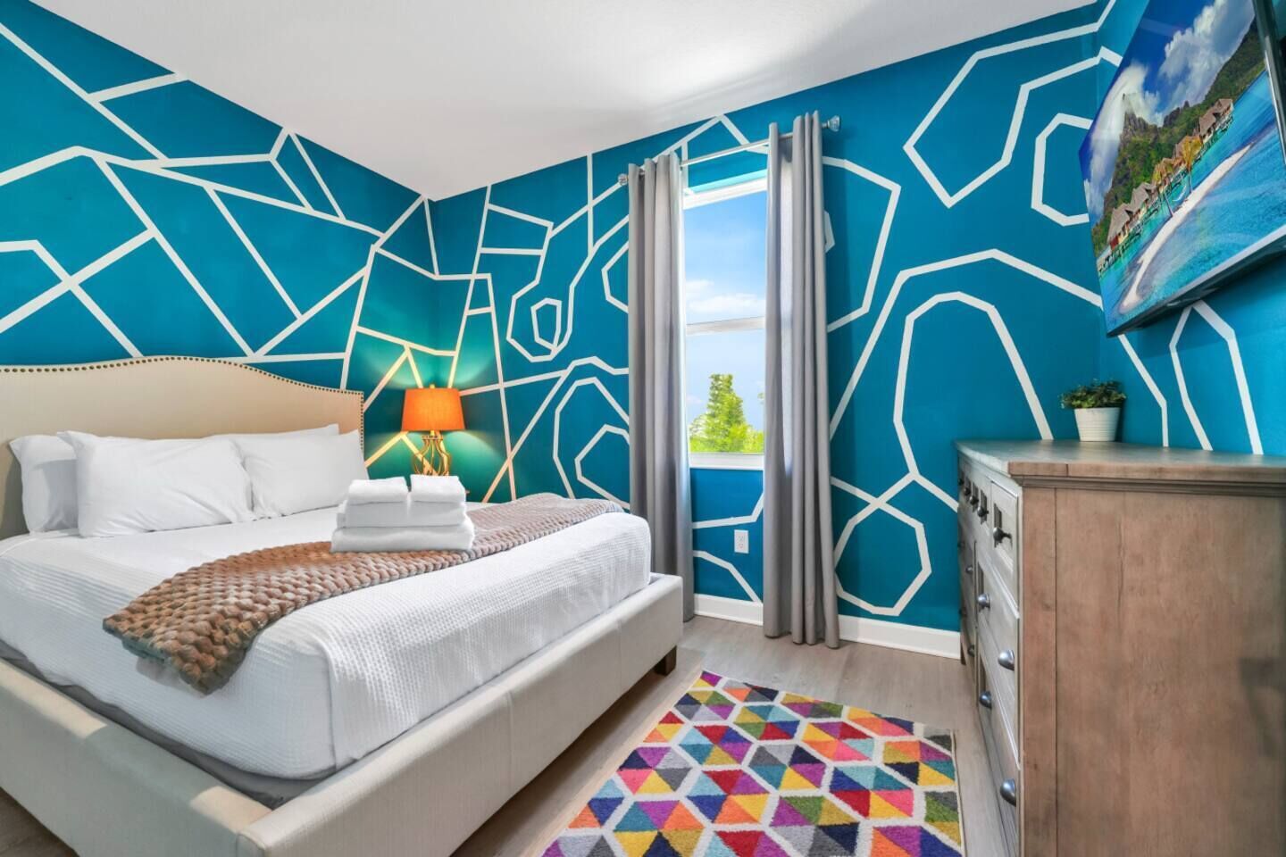 A hotel room with a bed, a nightstand, a lamp, a dresser, a colorful rug, and a large window with curtains.