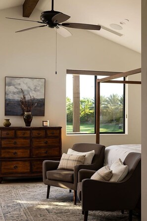 A room with a ceiling fan, a large window with a view of greenery, a wooden dresser, and a brown leather chair.