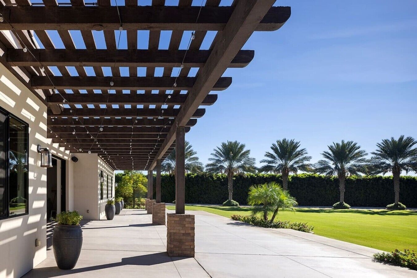 Un espacio exterior cubierto con un pérgola, plantas en macetas y un césped bien cuidado con palmeras.