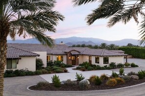 A two-story house with a tiled roof, surrounded by landscaped gardens and palm trees, with mountains in the background.