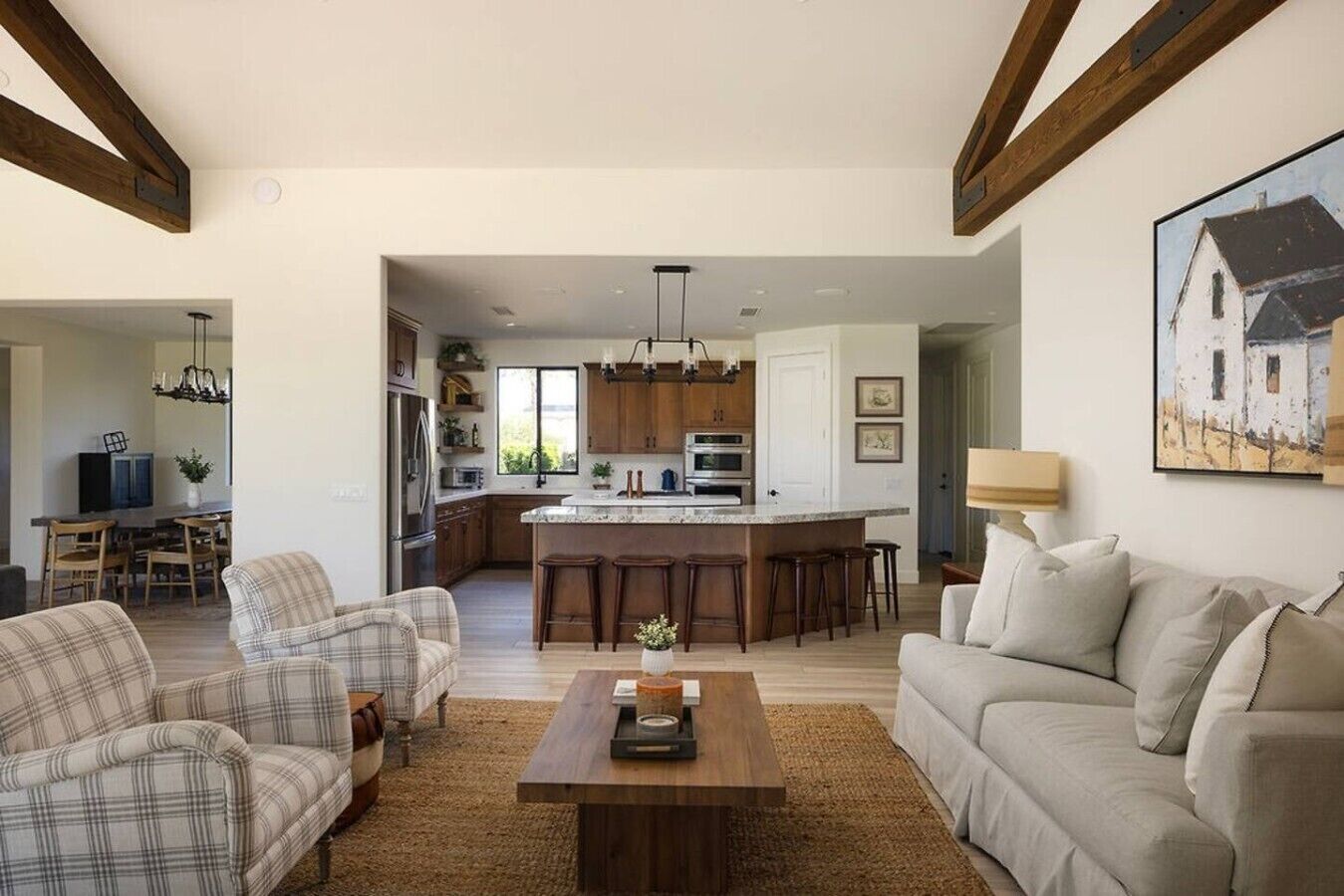Amplia sala de estar con una mesa de centro, un sofá y un comedor con sillas de madera. Se ve una cocina con una isla grande y taburetes de bar al fondo.