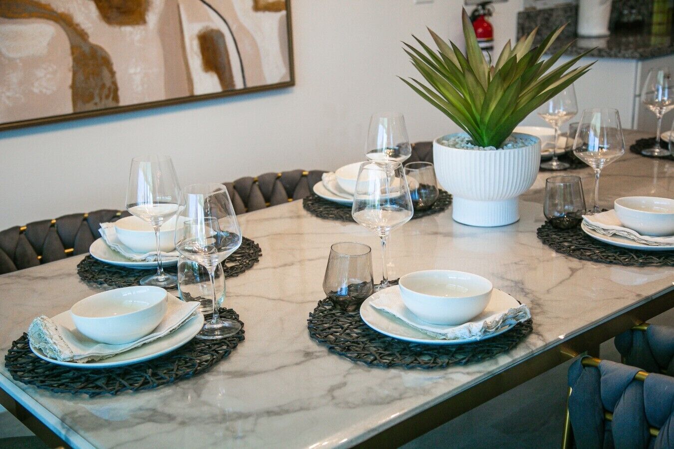 Mesa de jantar posta com pratos, tigelas e copos de vinho brancos, envolvida por jogos americanos de tecido escuro.