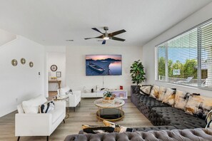 A modern living room with a white sofa, a dark tufted sofa, a round coffee table, and a television mounted on the wall. - 8BR Luxury Pool Spa Pickleball Hoops Game Room BBQ - Sleeps 28 (Coachella)