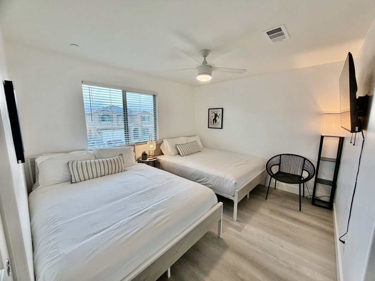 A bedroom with two beds, a ceiling fan, a window with blinds, a small table with a lamp, and a chair.