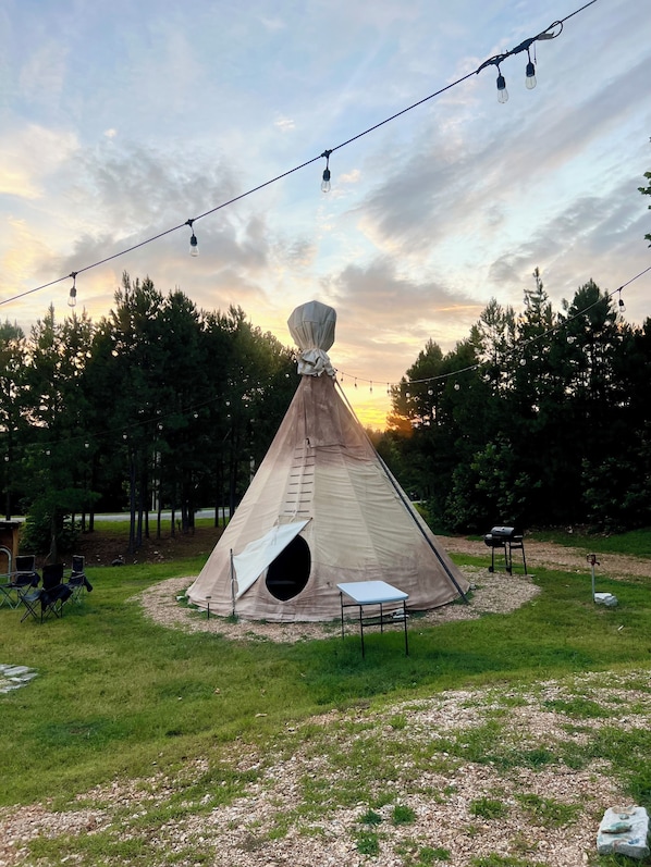 A teepee with a ladder, a table, and chairs in a grassy area with trees and string lights.