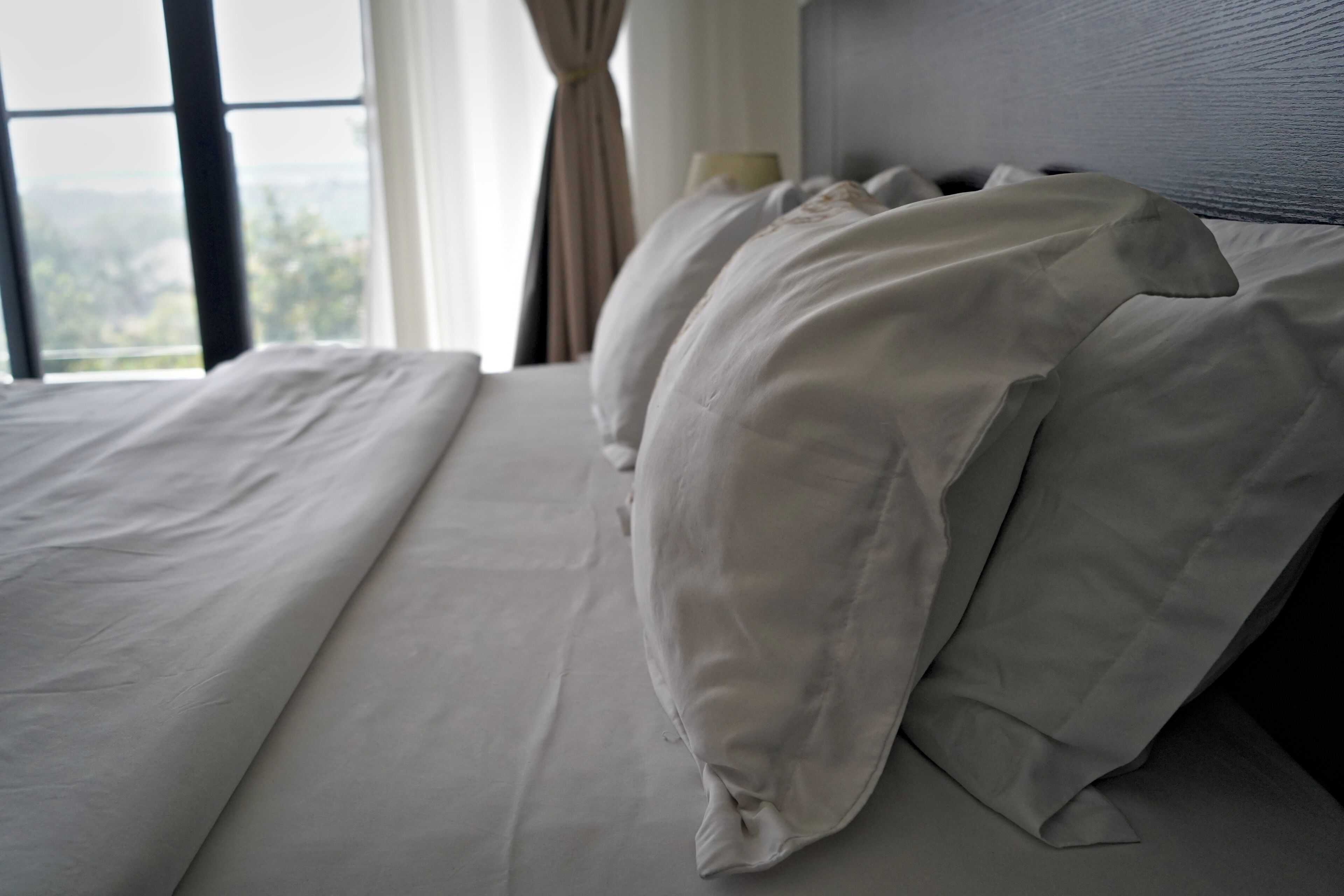 Un lit avec du linge de lit blanc et une tête de lit, une fenêtre avec des rideaux et une vue sur l’extérieur.