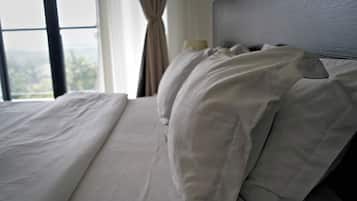 Un lit avec du linge de lit blanc et une tête de lit, une fenêtre avec des rideaux et une vue sur l’extérieur.
