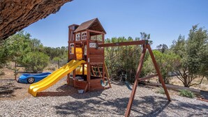 A wooden play structure with a slide and swings, set on gravel with trees in the background. - Casa Robles by Avantstay Spa + Firepit + BBQ Rural Eastside Paso Robles (Paso Robles)